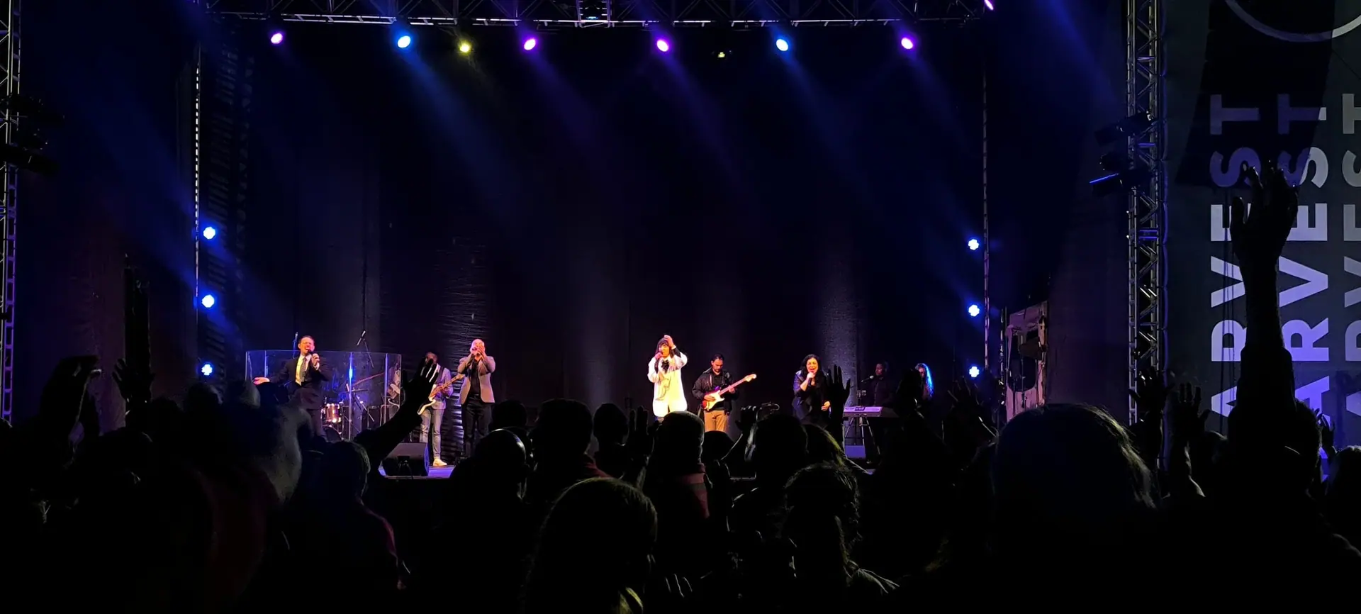A worship band on the CRC Harvest stage under blue and white production lighting, with the crowd in front
